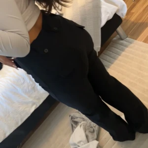 Svarta kostymbyxor med raka ben - Snygga svarta kostymbyxor med hög midja och raka ben. Byxorna har fickor med knappdetaljer bak och är perfekta för en clean och stilren look. Materialet känns mjukt och följsamt, vilket gör dem bekväma hela dagen.