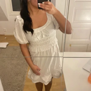 Vit kort klänning - Supersöt vit kort klänning med puffärmar, smockad midja och volangkant nedtill. Klänningen har en rundad halsringning och är gjord i mjuk bomull. Perfekt för dig som gillar romantisk och trendig stil.