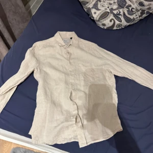 Beige linneskjorta från H&M Essentials - Säljer en stilren beige skjorta från H&M Essentials i linne. Skjortan har klassisk krage, långa ärmar med knappar vid manschetten och en avslappnad passform. Perfekt för en clean och enkel look. Denna passar även m