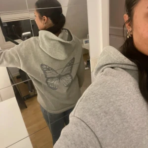 Grå hoodie med fjärilstryck - Säljer en grå hoodie med stor fjäril i lila på ryggen. Tröjan har huva, lång ärm och en loose passform. Mjuk och skön, perfekt för chill dagar. Materialet är bomull och polyesterblandning. Enkel design framtill och snygg detalj på baksidan.