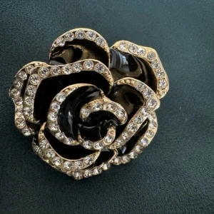 Svart och guld blomformad brosch - Unik brosch i form av en ros med svarta och guldfärgade detaljer, dekorerad med glittrande stenar längs kanterna. Baksidan är helgjuten i guldton och har nål för enkel fastsättning. Perfekt för att ge din outfit en extra touch av glamour.