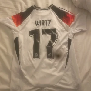 Tysklands Wirtz fotbollströja Adidas - Säljer en vit Tysklands fotbollströja från Adidas med Wirtz och nummer 17 på ryggen. Tröjan har svarta och röda detaljer på axlarna, tre svarta ränder på ärmarna och Tysklands landslagsmärke med fyra stjärnor på bröstet. Materialet är lätt och andas, perfekt för match eller träning.