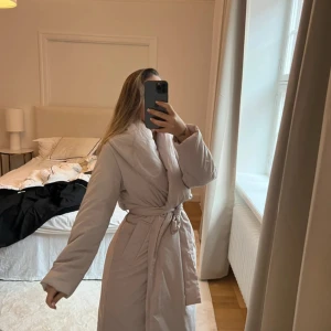 Beige lång dunjacka med bälte - Säljer en beige lång dunjacka med bred krage och bälte i midjan. Jackan har en omlott-design och stora fickor framtill. Perfekt för dig som vill ha en mysig och stilren look 