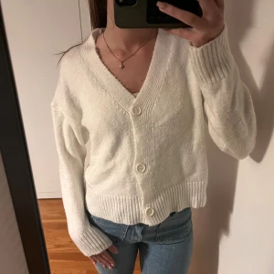 Vit stickad kofta med knappar - Mysig vit stickad kofta med v-ringning och stora vita knappar framtill. Koftan har långa ärmar och ribbade muddar vid ärmslut och nederkant. .