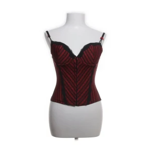 Randig vinröd och svart korsett - Säljer en snygg korsett i vinrött och svart med ränder och spetsdetaljer vid bysten. Axelbanden är justerbara och modellen är figurnära med markerad midja. Perfekt för dig som vill sticka ut med en edgy look.