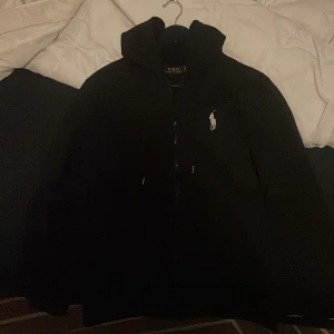 Svart hoodiejacka från Polo Ralph Lauren - Svart hoodiejacka från Polo Ralph Lauren med dragkedja och snörning i huvan. Klassisk vit Polo-logga på bröstet, två fickor fram och långärmad passform. Jackan är i mjukt bomullsmaterial och har en clean, stilren look som funkar till streetwear.