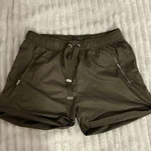 Träningsshorts  - Snygga olivgröna träningsshorts med elastisk midja och snörning framtill. Shortsen har dragkedjefickor på sidorna och är tillverkade i ett lätt, perforerat syntetmaterial som andas. Perfekta för sommarens alla aktiviteter.