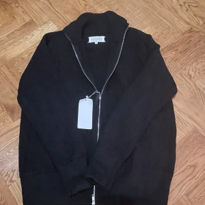 Svart stickad kofta från Maison Margiela - Svart ribbstickad kofta från Maison Margiela med hög krage och silverfärgad dragkedja. Tröjan har lång ärm och klassiska vita stygn i nacken. Perfekt för dig som gillar minimalistisk och stilren design.