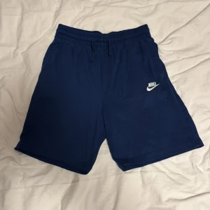 Marinblå shorts från Nike  - Snygga marinblå shorts från Nike med klassisk logga på vänster ben. Shortsen har elastisk midja med snörning och är tillverkade i mjuk bomull, perfekta för sommaren eller träning.