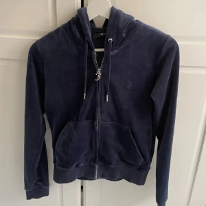 Mörkblå velour hoodie med dragkedja - Mjuk och skön mörkblå hoodie från Juicy Couture i velour med huva, snörning och dragkedja framtill. Tröjan har långa ärmar och en stor ficka på magen. 