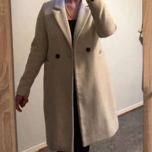 Beige kappa från Vero Moda - Stilren beige kappa från Vero Moda med klassisk krage och två svarta knappar framtill. Långärmad modell med rak passform och diskreta fickor på sidorna. Perfekt för dig som gillar en enkel och tidlös look.