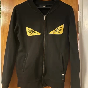 Svart hoodie från Fendi med ögon - Svart hoodie från Fendi med dragkedja framtill och två fickor. På bröstet finns två gula applikationer som ser ut som ögon med svarta blixtdetaljer. Tröjan har lång ärm och en stilren, modern look som sticker ut.