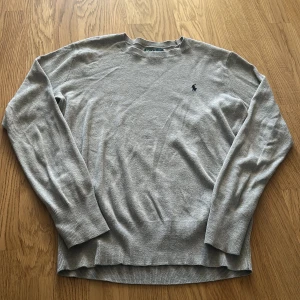 Grå tröja från Polo Ralph Lauren - Snygg grå tröja från Polo Ralph Lauren med klassisk rund hals och långärmad design. Tröjan har det ikoniska mörkblå Polo-logot broderat på bröstet och är tillverkad i mjuk bomull. Perfekt för dig som gillar stilrena och tidlösa plagg.