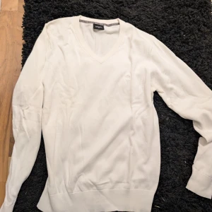 Vit v-ringad tröja från Adam stl M - Vit v-ringad tröja från Adam i mjuk bomull. Klassisk design med ribbad kant vid hals, ärmslut och nederkant. Diskret broderad logga på bröstet. Perfekt för dig som gillar stilrena och enkla plagg.