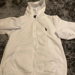 Vit kofta från Polo Ralph Lauren - Vit hoodie från Polo Ralph Lauren med dragkedja och huva. Tröjan har lång ärm, ribbade muddar och klassisk Polo-logga broderad på bröstet. Tillverkad i mjuk bomull med stretch för extra komfort. Perfekt för chill dagar och streetwear.
