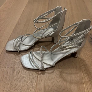 Silvriga klackar - Snygga silvriga klackar med smala remmar dekorerade med strass. Skorna har fyrkantig tå, öppen tå och en låg klack. Perfekta för dig som vill sticka ut med glittriga detaljer och elegant design. Från Dream Paris och använda en gång, nypris var mellan 400-500kr