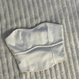 Vit korsett-topp från H&M XS - Säljer en vit korsett-topp från H&M i storlek XS. Toppen har en stilren bandeauform med markerade sömmar som ger en snygg siluett. Materialet är mjukt och stretchigt, perfekt för att skapa en cool look. Passar dig som gillar minimalistisk stil.