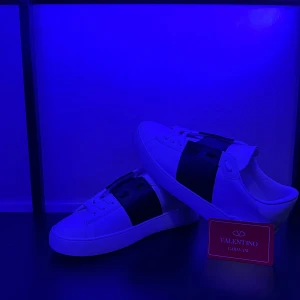 Valentino Garavani Open sneakers vit/svart - Snygga Valentino Garavani Open sneakers i vitt skinn med bred svart rem över mitten. Klassisk rund tå, platt sula och diskret logga på plösen och sulan. Perfekta för dig som vill ha en clean men lyxig look. Kommer med originalpåse.