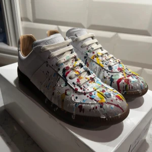 Maison Margiela sneakers paint splatter - Unika sneakers från Maison Margiela i vitt skinn med färgstänk i rött, gult, blått och svart. Klassisk låg modell med snörning, bruna gummisulor och rund tå. Skorna har en konstnärlig vibe och sticker ut med sitt färgglada mönster. Skokartong ingår ej