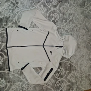 Nike Tech tracksuit  - Nike Tech Fleece set med hoodie och matchande byxor i ljusvit färg med svarta detaljer. Hoodien har dragkedja, fickor och en snygg svart Nike-logga på bröstet. Byxorna har snörning i midjan och logga på benet. Materialet är mjukt och skönt fleece, perfekt för chill eller träning.