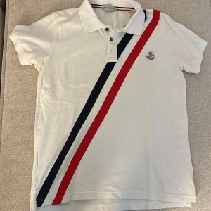 Vit Moncler piké med röd och marinblå rand - Vit pikétröja från Moncler med klassisk krage och knappar framtill. Snygga diagonala ränder i rött och marinblått över framsidan samt Moncler-logga på bröstet. Kortärmad och tillverkad i mjuk bomull