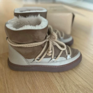 Beige vinterboots från Inuikii stl 41 - Säljer ett par snygga classic vinterboots från Inuikii i beige och ljusbrunt skinn och mocka. Skorna har grova snören, rund tå och är fodrade med mjuk päls för extra värme. Perfekta för kalla dagar och har en chunky brun sula som ger cool stil.Nypris 3100kr
