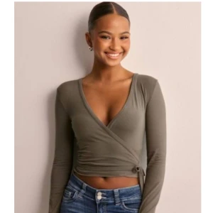 Olivgrön omlottopp med knyt från Nelly - Snygg olivgrön omlottopp med lång ärm och djup v-ringning. Toppen är croppad och har ett knyt i sidan som ger en cool detalj. Perfekt att styla med jeans eller kjol för en trendig look. Ny pris 299kr