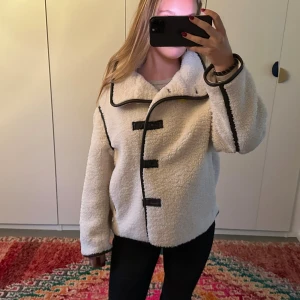 Vit teddyjacka med svarta detaljer - Mysig vit teddyjacka med svarta kantband och stora knappar framtill. Jättefint skick o passar till allt