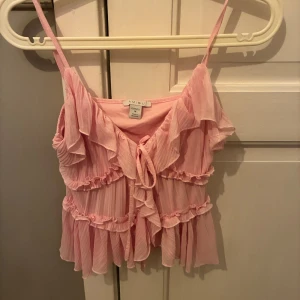 Rosa volangtop från Amisu - Söt rosa linnetopp från Amisu med tunna axelband och flera volanglager. Toppen har knytning framtill och är gjord i ett lätt, transparent syntetmaterial med rynkade detaljer. Perfekt för fest eller sommar! Köparen står för frakten!