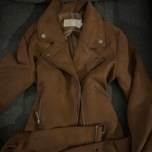 Brun mocka bikerjacka NLY Trend - Snygg jacka i brun mocka från NLY Trend, med coola silvriga dragkedjor och bälte i midjan. Jackan har klassisk krage med tryckknappar och två fickor med dragkedja. 