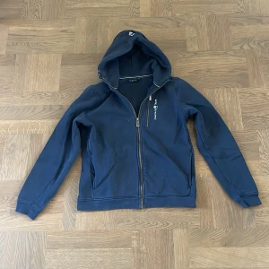 Marinblå hoodie från Sail Racing - Snygg marinblå hoodie från Sail Racing med dragkedja framtill och broderad logga på bröstet. Tröjan har huva, långa ärmar och är tillverkad i mjuk bomull med fleece på insidan. Använd bra mängd men fortfarande väl omhändertagen. Perfekt för hösten och vintern, storlek: 170.