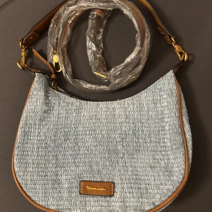 Blå axelväska från Tamaris - Snygg axelväska från Tamaris i ljusblått tyg med bruna skinn-detaljer och justerbar axelrem. Väskan har en rundad form och ett litet Tamaris-märke i brunt skinn framtill. Aldrig använd men köpt online så har inte förpackningen kvar.