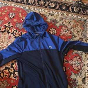 Blå vindjacka från Lacoste Sport - Säljer en snygg tvåfärgad vindjacka från Lacoste Sport i marinblått och blått. Jackan har huva, dragkedja hela vägen och den klassiska krokodilloggan på bröstet. Perfekt för dig som gillar sportig stil och vill ha något lätt och funktionellt.