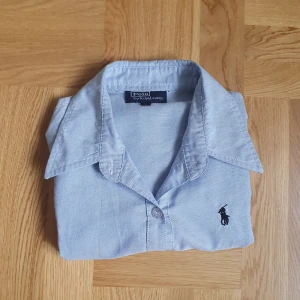 Ljusblå skjorta från Polo Ralph Lauren - Snygg ljusblå skjorta från Polo Ralph Lauren med klassisk krage och lång ärm. Skjortan har knappar framtill och det ikoniska Polo-logot broderat på bröstet. Tillverkad i mjuk bomull som känns skön mot huden. Perfekt för dig som gillar stilrena och tidlösa plagg. Skriv vid minsta fråga eller fundering! 