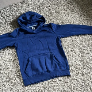 Blå hoodie från Blue Ridge Denim - Snygg marinblå hoodie från Blue Ridge Denim med klassisk känguruficka och huva. Tröjan har diskret logga på ärmen och en liten BR-detalj vid mudden. Perfekt för dig som gillar en enkel och stilren look.