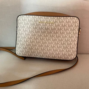 Michael Kors axelväska  - Snygg axelväska från Michael Kors i beige och brunt med MK-logga över hela väskan. Väskan har guldfärgad dragkedja, justerbar rem och flera fack inuti. Tillverkad i syntetmaterial med detaljer i skinn. 