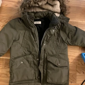 Olivgrön jacka med fuskpäls stl 116, 5-6 år - Parkajacka från H&M i olivgrönt med stor huva och fuskpäls. Jackan har två stora fickor med lock och knappar, samt dragkedja framtill. Fodrad insida och ribbade muddar vid ärmslut. Perfekt för kalla dagar. Obs dragkedjan defekt ,  behöver bytas ut 