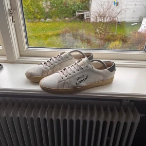 Saint Laurent vita sneakers i skinn - Snygga vita sneakers från Saint Laurent med handskriven logga på sidan. Skorna har beige sula, svarta detaljer vid hälen och är tillverkade i skinn. Klassisk låg modell med snörning och rund tå, perfekt för en clean och stilren look. | Ny pris: 7116kr | Vårt pris: 2200kr! | 🙌💯| Priset kan diskuteras! |