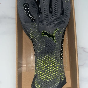 Puma målvaktshandskar svart/grå - Snygga målvaktshandskar från Puma i svart och grått med gula detaljer. Ovansidan är stickad med gula inslag och Puma-logga, medan handflatan är svart med vita prickar för extra grepp. Perfekta för fotbollsträning eller match.