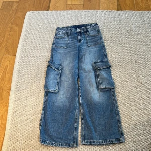 Blå wide leg cargojeans från H&M - Säljer ett par blå cargojeans från H&M med breda ben och låg midja. Jeansen har justerbar midja och stora fickor på sidorna. Perfekta för en avslappnad och trendig look. Materialet är klassisk denim.