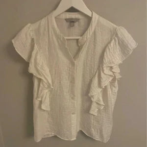 Vit blus med volanger från H&M XS - Supersöt vit blus från H&M i storlek XS. Blusen har korta ärmar med snygga volangdetaljer längs axlarna och knappar framtill. Materialet är lätt och luftigt, perfekt för varmare dagar. Enkel att styla med jeans eller kjol för en fräsch look.