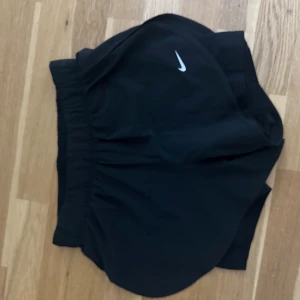 Svarta Nike Dri-FIT träningsshorts - Svarta träningsshorts från Nike med Dri-FIT-teknologi. De har elastisk midja, rundade benkanter och en liten vit Nike-logga framtill. Perfekta för löpning eller gym, tillverkade i lätt och snabbtorkande syntetmaterial.