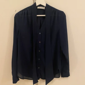 Mörkblå skjortblus från Carin Wester  - Snygg mörkblå skjortblus från Carin Wester med knappar framtill och långa ärmar. Skjortan har en cool knyt-detalj vid halsen som ger extra stilpoäng. Tillverkad i ett lätt och luftigt material som känns skönt mot huden. Perfekt för dig som gillar en clean och modern look.