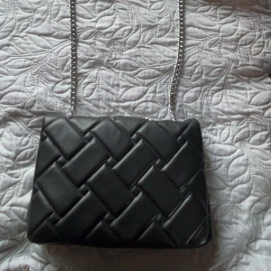 Svart quiltad axelväska med kedja - Snygg svart axelväska med quiltad design och breda, vadderade rutor. Väskan har en silverfärgad kedjerem med läderdetalj upptill för extra komfort. Perfekt storlek för det viktigaste och stängs med magnetlås. Stilren och trendig look.