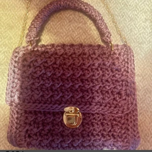 Lila stickad handväska med guldkedja - Superfin  handmade lila handväska i stickat material med snyggt flätmönster. Väskan har ett stadigt handtag, guldfärgad kedja och ett coolt guldfärgat spänne framtill. Perfekt accessoar för att lyfta din outfit med en unik touch.