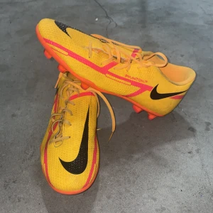 Nike Phantom GT fotbollsskor gula - Säljer ett par Nike Phantom GT fotbollsskor i en riktigt snygg gul färg med svarta och rosa detaljer. Skorna har snörning, platt sula med dobbar och är tillverkade i syntetmaterial för bästa grepp på planen. Perfekta för dig som vill sticka ut på matchen!