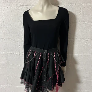 Svart kjol med rosa detaljer och kedjor storlek L. - Unik svart kjol med rosa band, kedjor och korsdetaljer. Asymmetrisk kant med svarta nätpartier och långa rosa snören som hänger ner. Kjolen har flera metallringar upptill och edgy goth-vibe. Perfekt för dig som gillar alternativ stil. Midjevidd 84 cm.