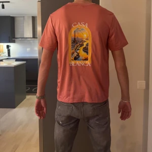 Casa Blanca korallrosa t-shirt - Snygg korallrosa t-shirt från Casa Blanca med tryck av stadsmotiv och solnedgång på ryggen samt mindre tryck på bröstet. Klassisk passform med rund hals och korta ärmar. Perfekt för dig som vill sticka ut med färg och coolt print.