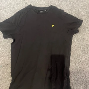 Mörkgrå t-shirt från Lyle & Scott - Mörkgrå t-shirt från Lyle & Scott med klassisk rund hals och korta ärmar. På bröstet sitter den ikoniska gula örnloggan. Tillverkad i mjuk bomull för en skön känsla och enkel stil.