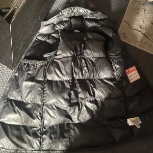 Svart jacka från Nike Therma-Fit - Svart jacka från Nike med Therma-Fit-teknologi. Jackan har quiltad design, diskret Nike-logga på bröstet och dragkedja framtill. Perfekt för kalla dagar när du vill hålla värmen men ändå vara rörlig. Passar dig som gillar sportig och clean stil.